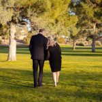 A couple walking arm-in-arm in a sunny park, surrounded by trees and grass.
