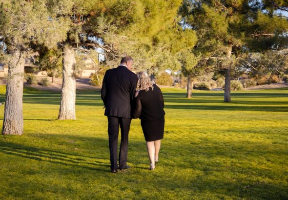 A couple walking arm-in-arm in a sunny park, surrounded by trees and grass.